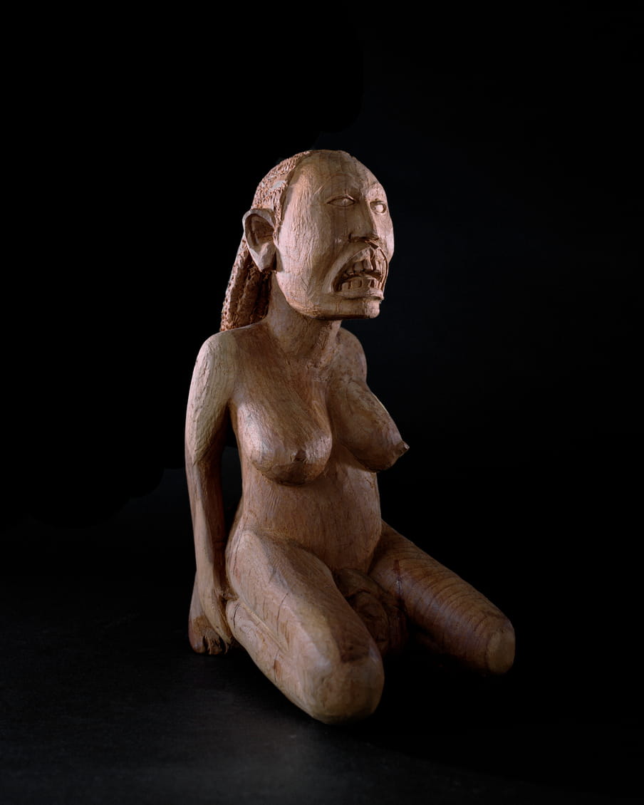 Photo of a wooden statue of a female figure giving birth on her knees, her mouth is open, showing teeth - on a black background. Compared to the last image her proportions are more realistic.