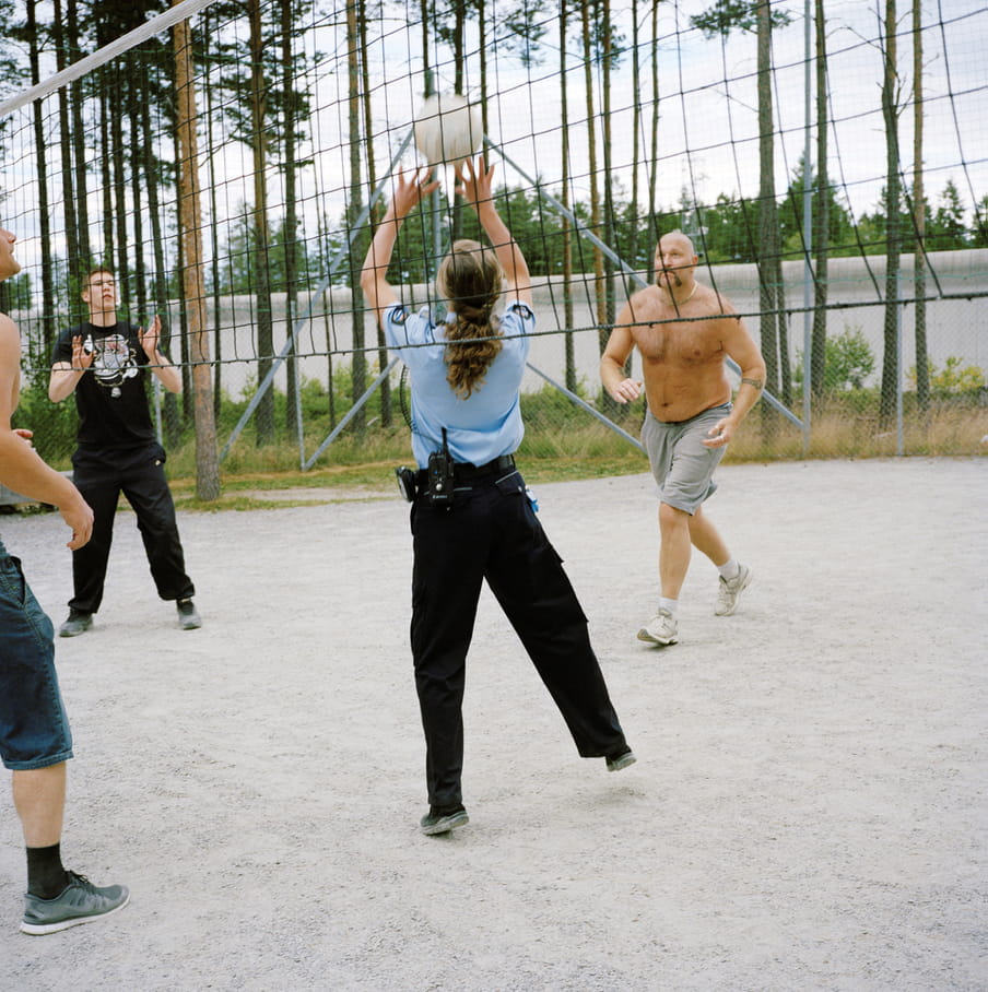 Four figures playing volleyball, central figure wears a prison guard outfit and jumps to reach the ball, the net runs through the picture. To the left is a figure in black with hands rising up, to the right a topless man in shorts and trainers running towards the ball