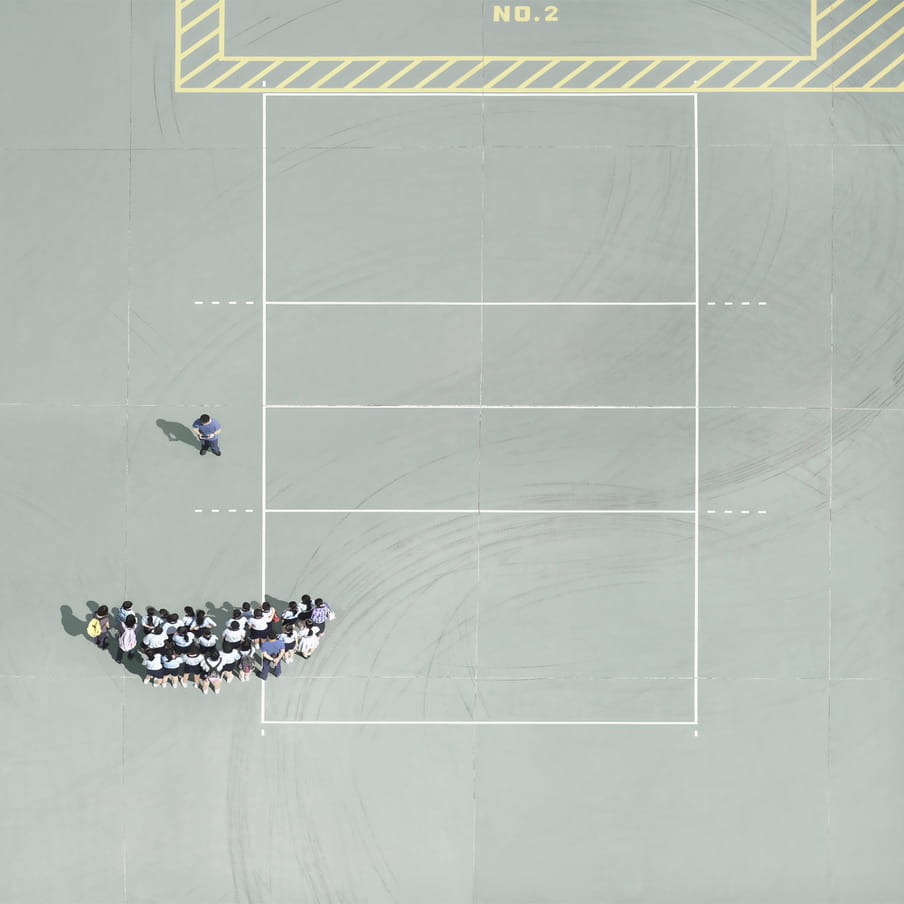 Aereal photograph on what looks like a mint green clay court with white and yellow box markings in the ground. To the bottom centre left, we see the heads and backs of a group of people gathered in a semi-circle, to have their picture taken by a photographer.