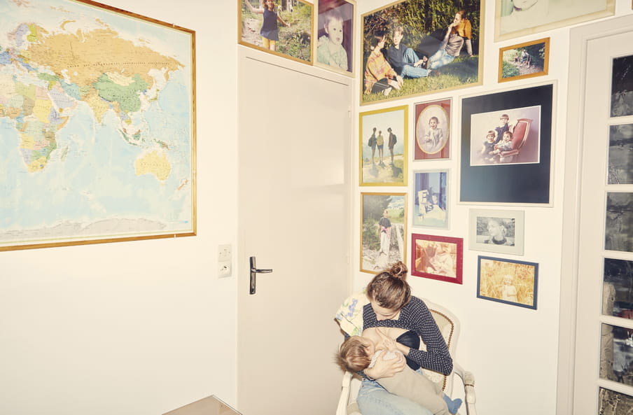 Photo of a room; on the left wall, a world map. A door in the middle with big photos above. On the right wall, 10 framed photos of different sizes featuring portraits of figures. On a chair lower right, a figure with brown hair wrapped in a bun on their head, with a towel over their left shoulder, holding their left breast as they feed a baby.