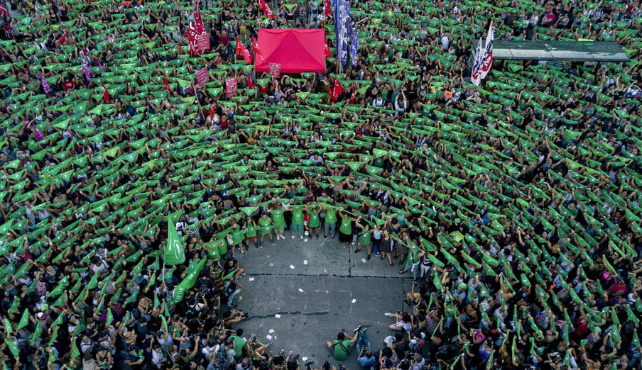 Photo of a crowd of people from above, all holding up a green bandana. There’s a hole in the crowd at the bottom, where photographers sit