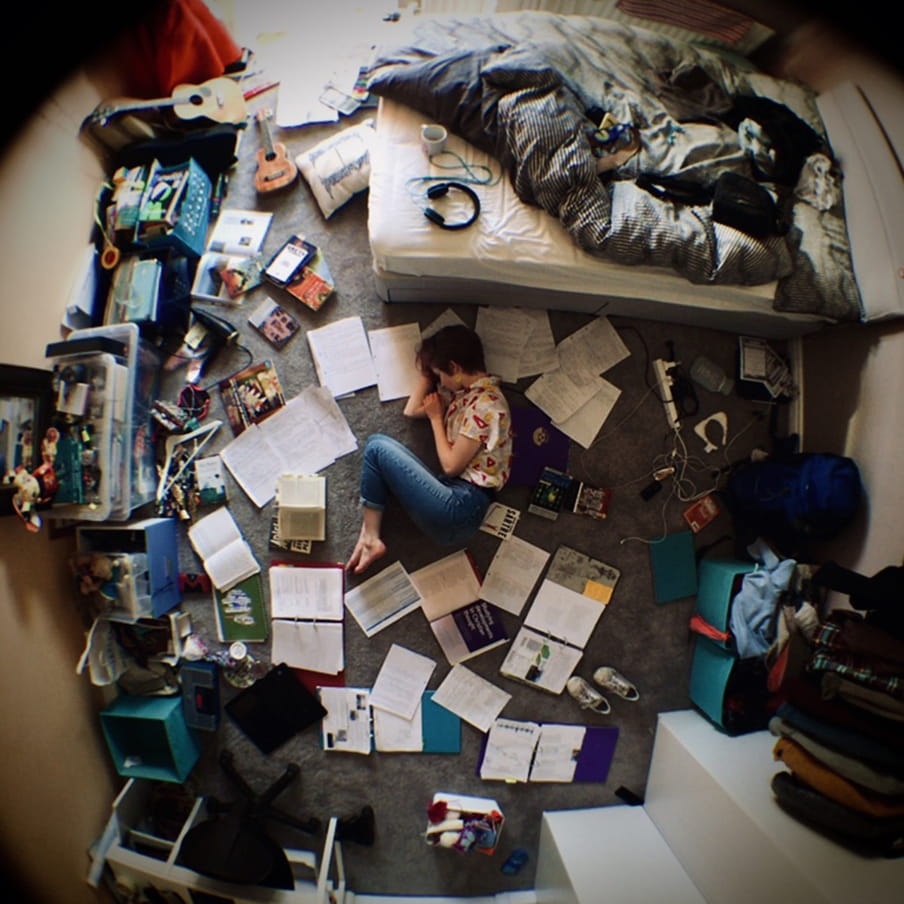 A boy is photographed curled up in the middle of his bedroom with many books and papers around him.