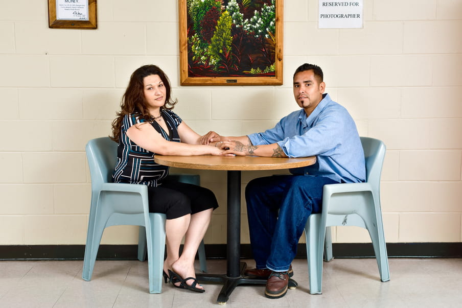 Photo of a man and a woman sitting on plastic chairs in front of a light yellow wall, a table in between them. The woman is wearing a blue, white and black shirt and black pants. The man is wearing hun prison uniform, jeans and a blue shirt. They are holding hands across the table.