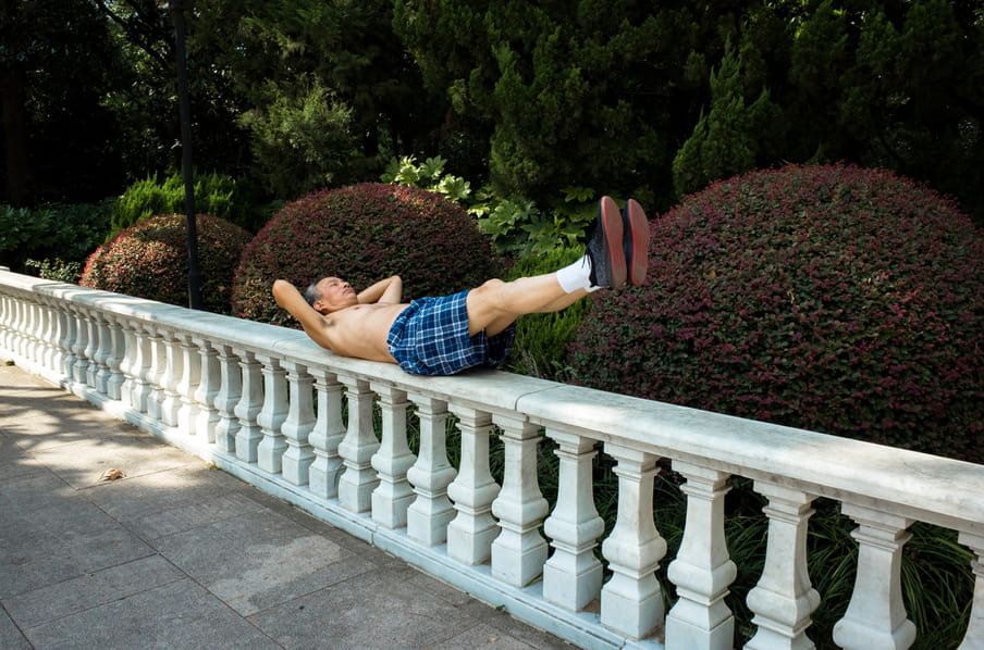 An old man lying on a stone wall in a park, doing stretching exercises.