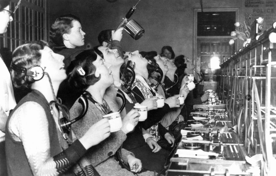 Black and white photograph of women operating telephone switchboards holding a cup and gargling with their head tilted backwards.