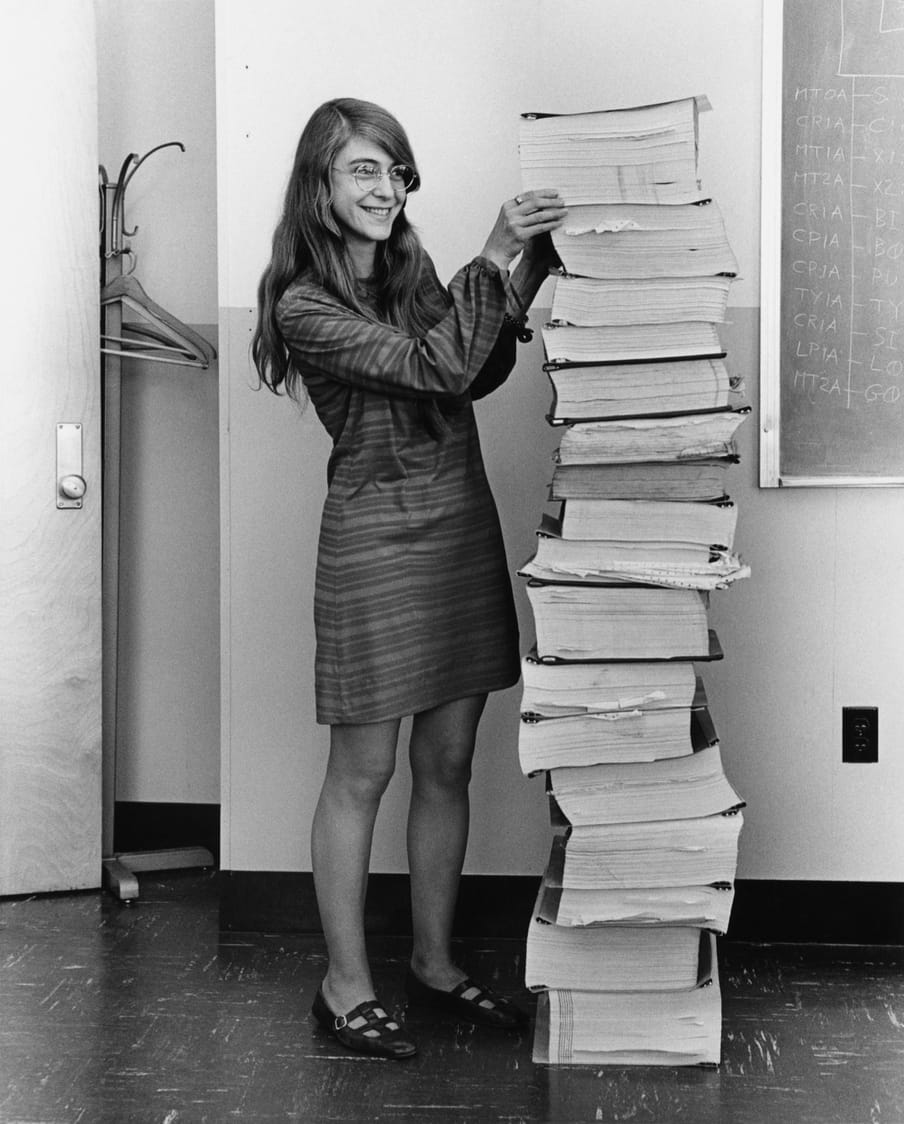 Black and white photograph of a woman wearing glasses, smiling, standing next to a pile of document as tall as her.