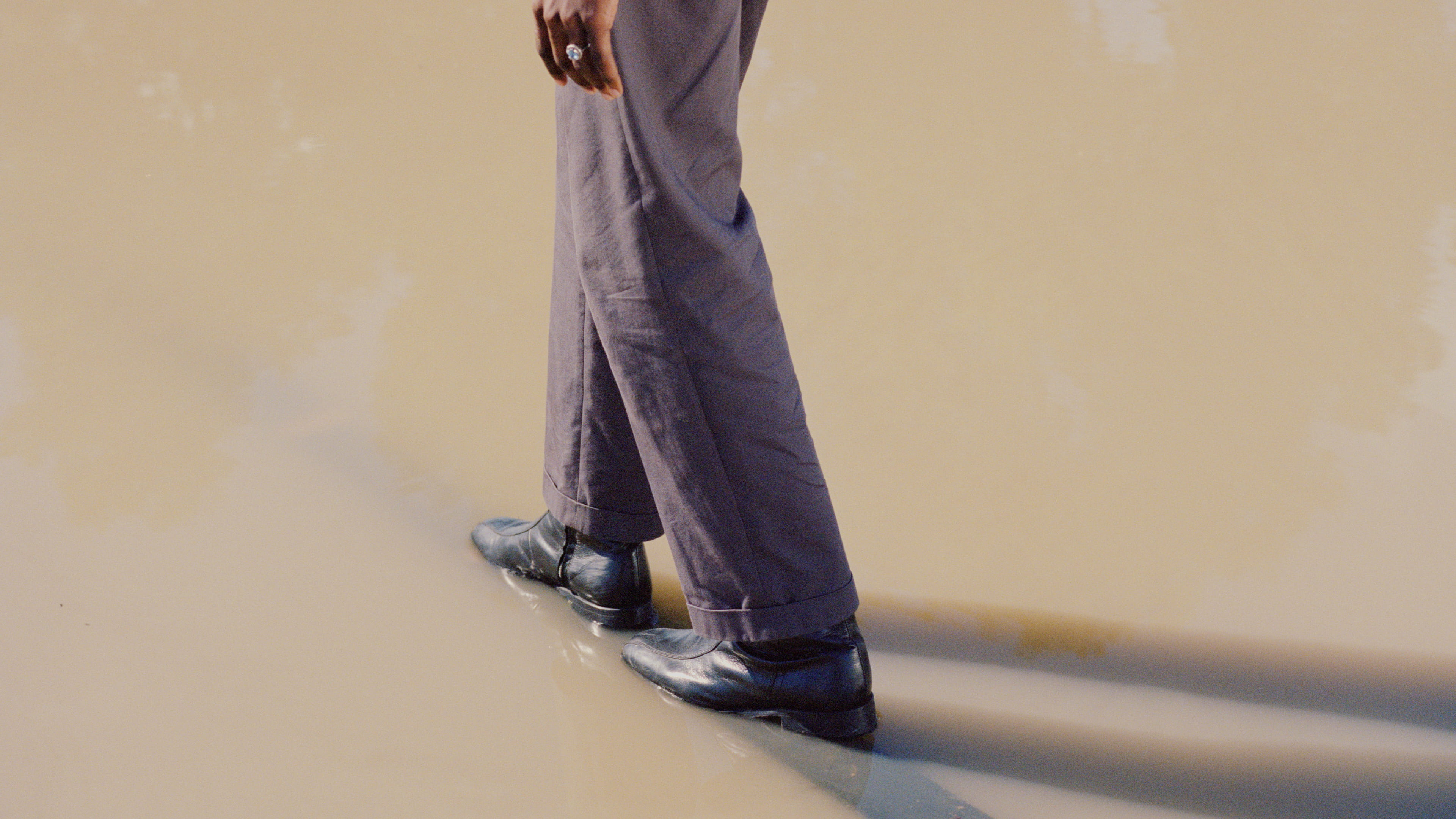 Photo of a person’s legs and black shoes, wearing baggy purple suit trousers, and the fingertips, walking through a pool of muddy water