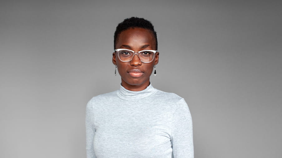 Photographic portrait of a woman with glassed, a turtleneck sweater and earrings - OluTimehin Adegbeye - against a grey background.