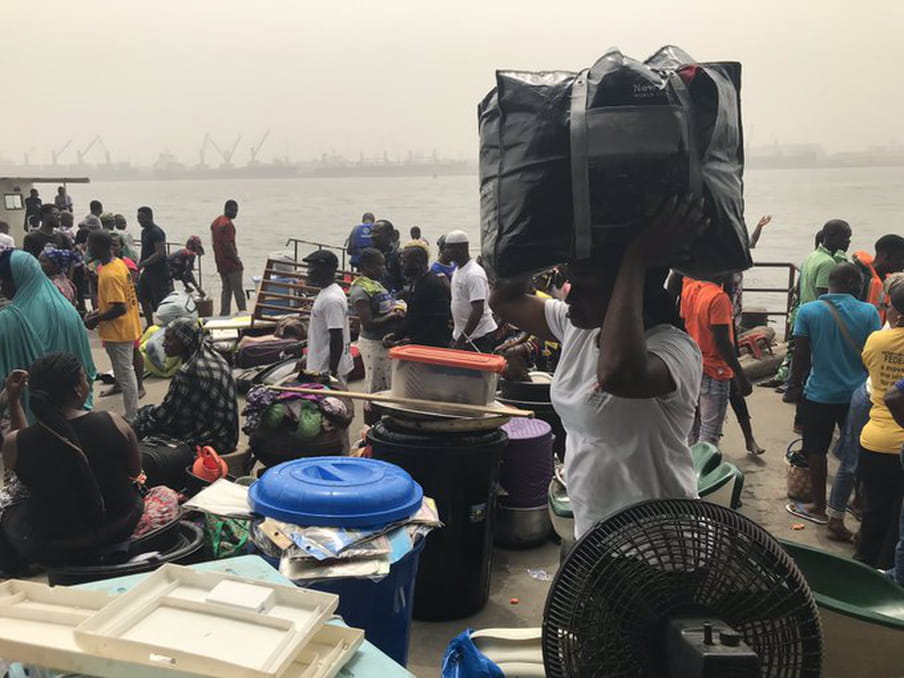 A woman carries a heavy black bag on her head. She is surrounded by people and their belongings. The lagoon is in the background.