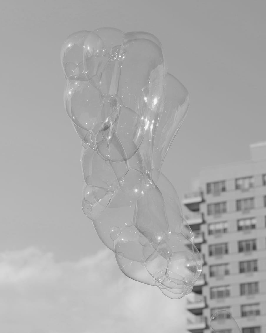 Black and white picture of a cloudy sky, with a high rise of flats in the background centre right, and a big stack of soapy bubbles stuck to one another, rising into the air, looking like a bunch of balloons