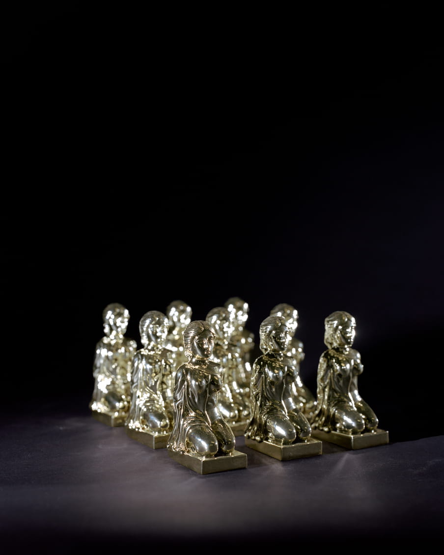 Photo of nine similar silver statues of a woman sitting on her knees with her hands on her back, compared to the previous image there are more statues but they are also smaller.