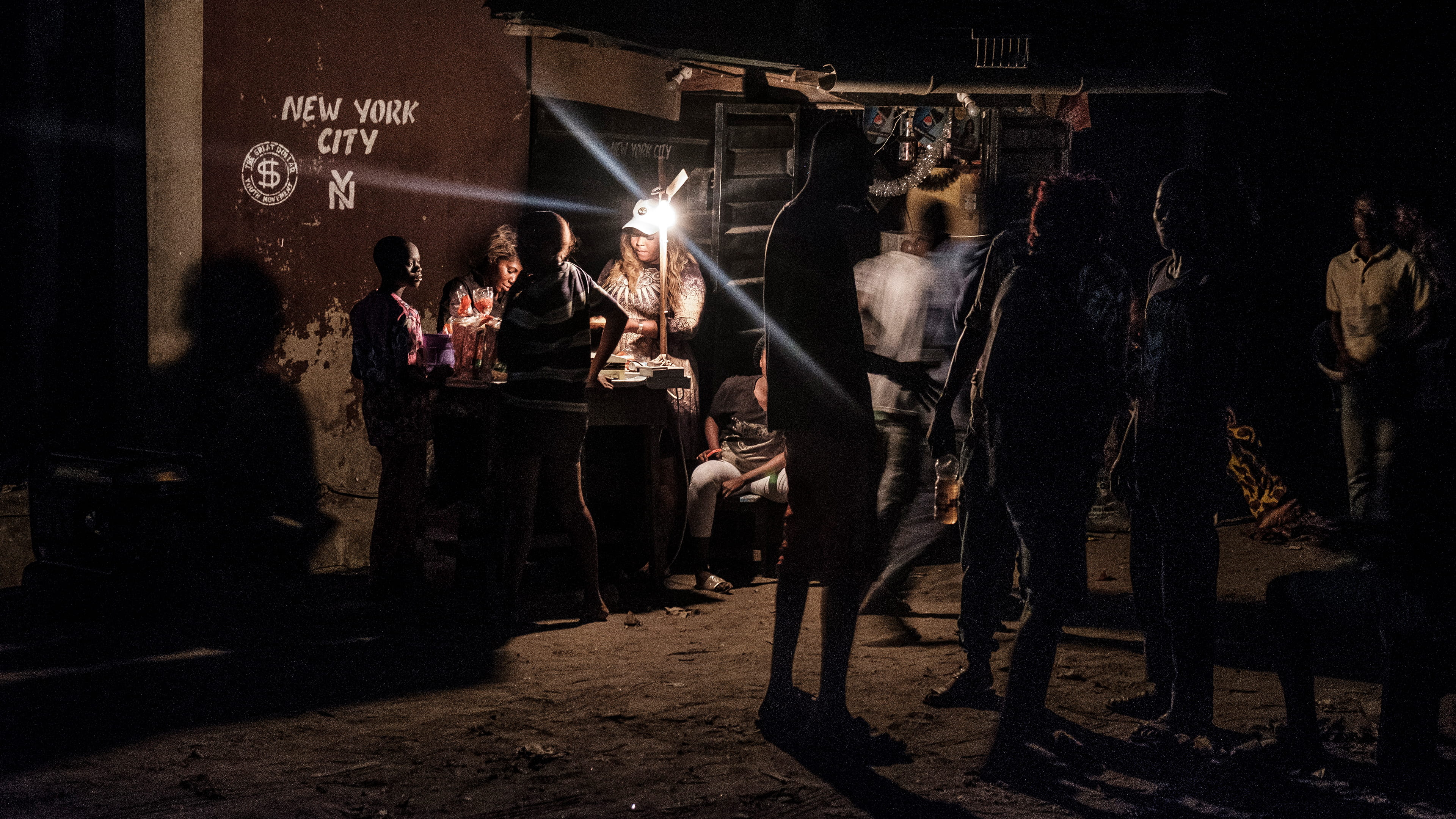 Photograph taken at night with people gathered together or standing around a small counter lit by a precarious lamp. The ground is sandy and on the brown wall in the back, the words New York city with a logo are painted in white.