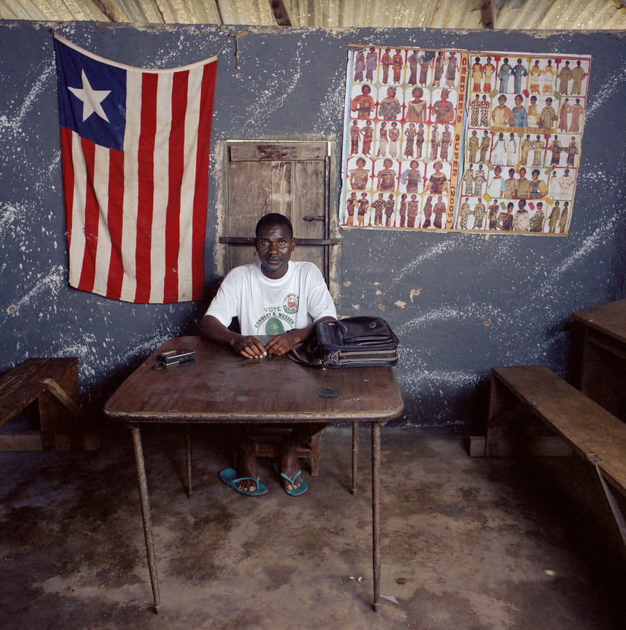 A Liberian male administrator sits at a sparse, dark brown desk. He’s wearing a t-shirt with a vote slogan and turquoise flip flops. There’s a leather bag on the desk. A flag is fastened to the wall behind him. Wooden tables and benches surround both sides of the desk.