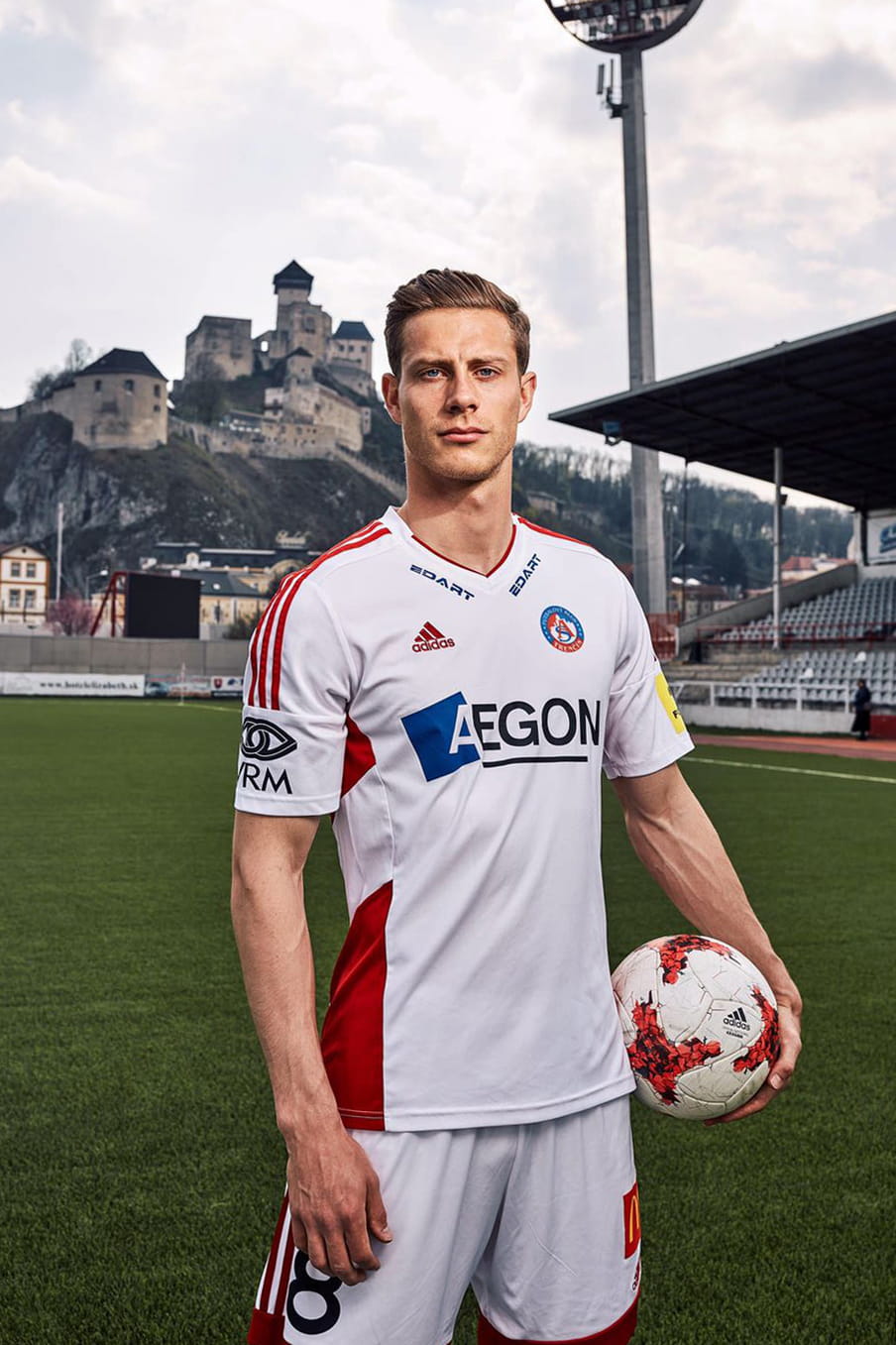 James Lawrence on the pitch of Slovenian football  club Tencin, posing for the camera with a ball loosely in his left hand. In the background a small castle on a hill top can be seen overlooking the stadium.