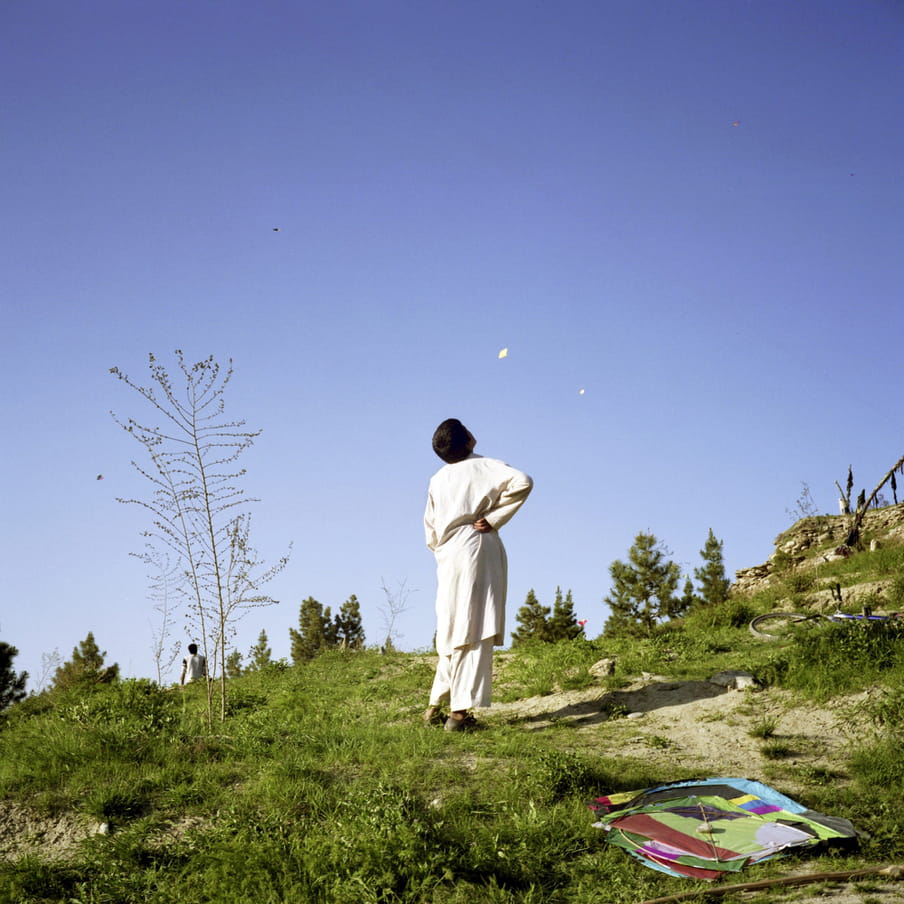  A boy in white traditional Afghan dress standing next to a kite lying on the ground, he is staring at the sky where several kites are flying