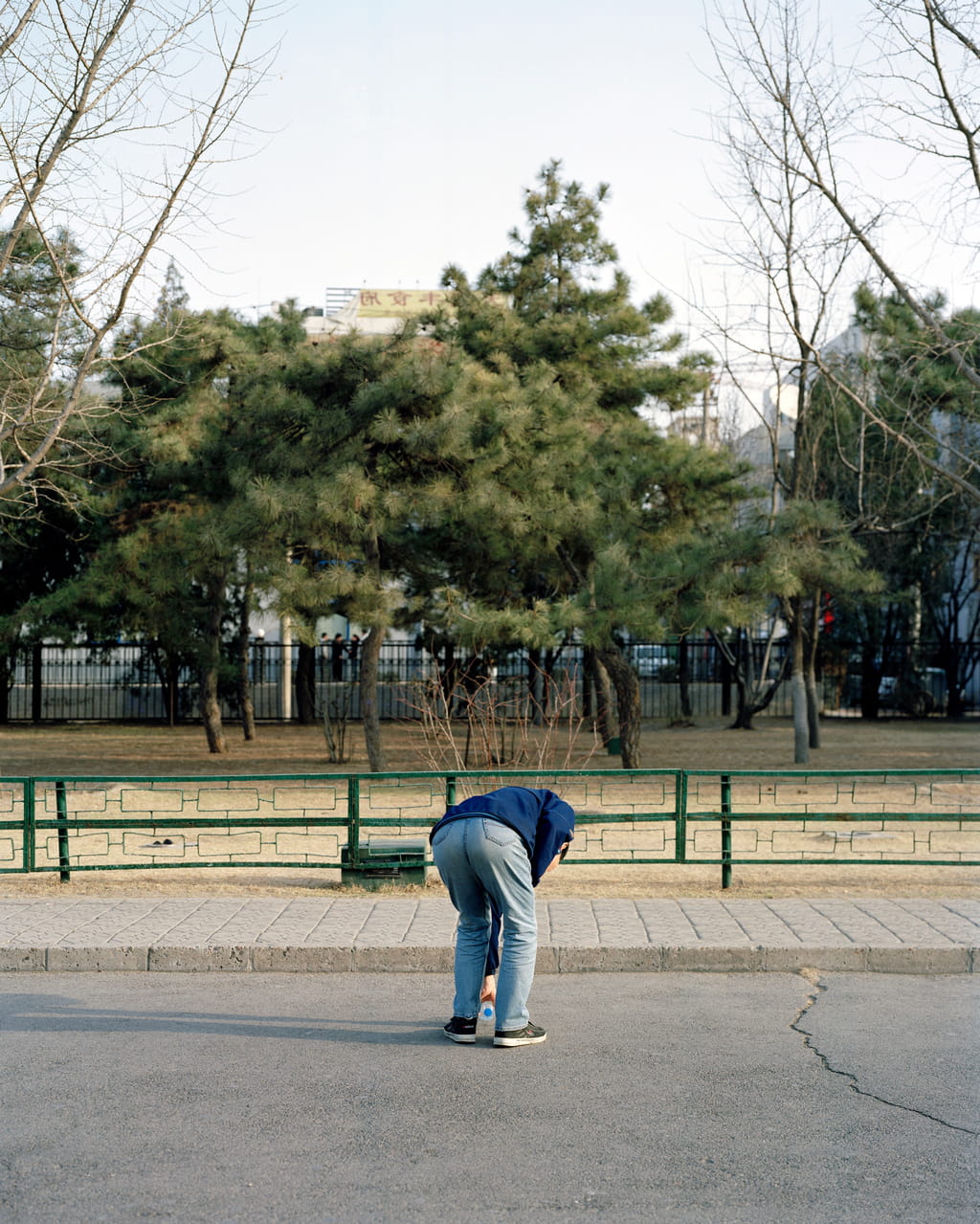 Photograph of a person picking up something that looks like a bottle from the road. In the background there is a small park.