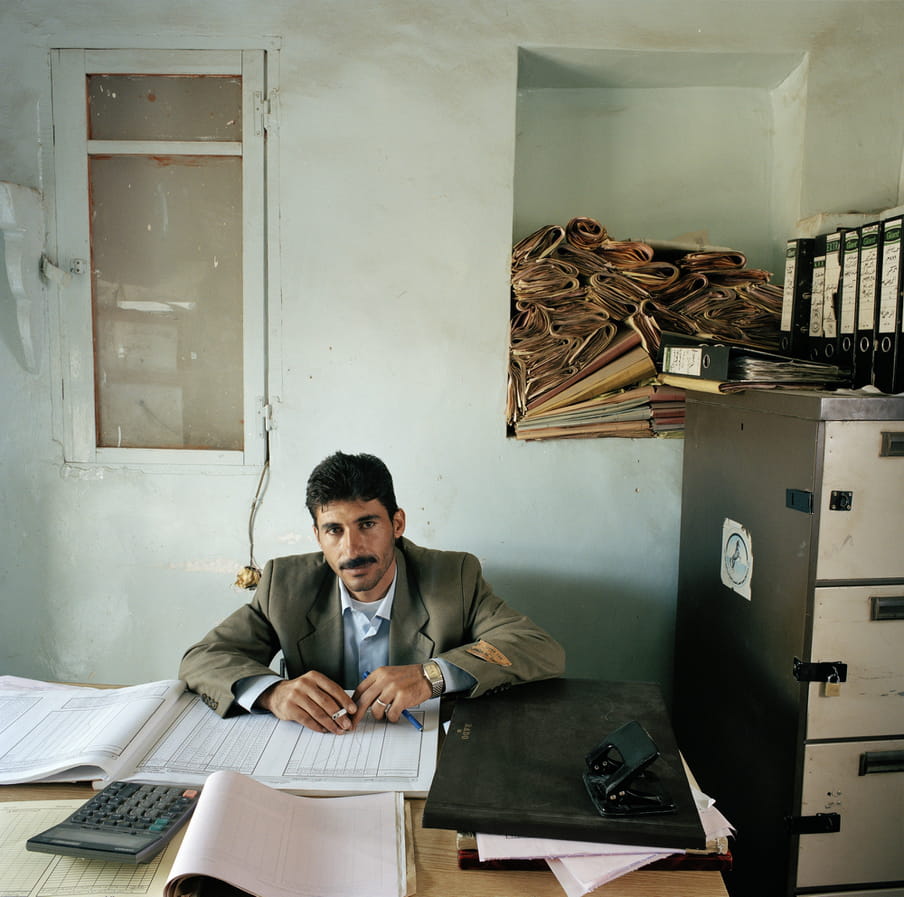 Male administrator in Yemen sitting at his desk covered with paperwork and a calculator. He holds a pen and a cigarette. He has dark hair and a moustache and wears a watch, a khaki-coloured jacket with a light-brown shirt underneath. There’s a filing cabinet next to the desk with big folders stacked on top. There’s a large niche in the wall in the right corner of the room, filled to the brim with brownish stacks of papers and files.
