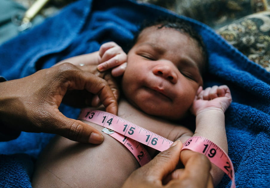 A newborn lies on a blue blanket with a patterned blanket under that, and has their hands up by their face; two hands are measuring the baby’s chest with a pinkish red measuring tape