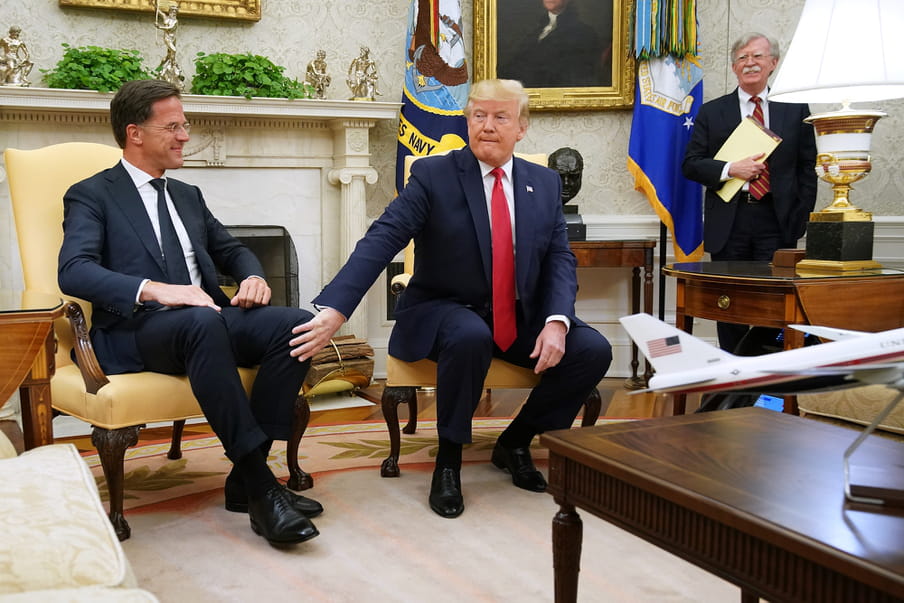 Two men wearing suits are sitting in yellow chairs. One man is touching the other man knee. The man who is touched is smiling. In the back, a third man is standing, also smiling. 