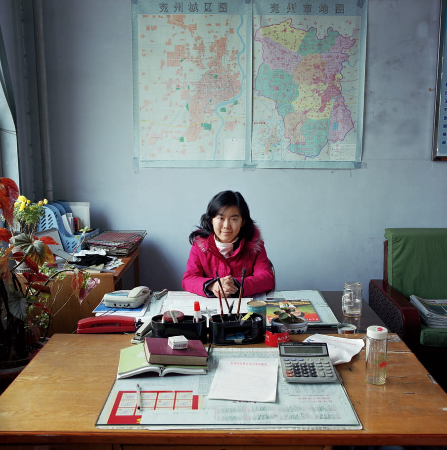 Female, smiling liaison officer sitting at a large desk. The desk has a calculator, some books, a landline and office supplies on it. The woman is wearing a thick, magenta winter jacket. On the left hand side we see a plant in front of the window. The wall is adorned with a Chinese map. At the back there’s a green armchair and another desk is being used as storage for files and a tray with paperwork.