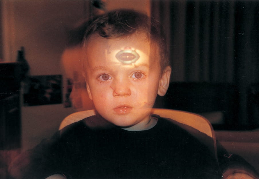 Photograph of a young boy, moving (out of focus), with the reflection of an UFO on his forehead.