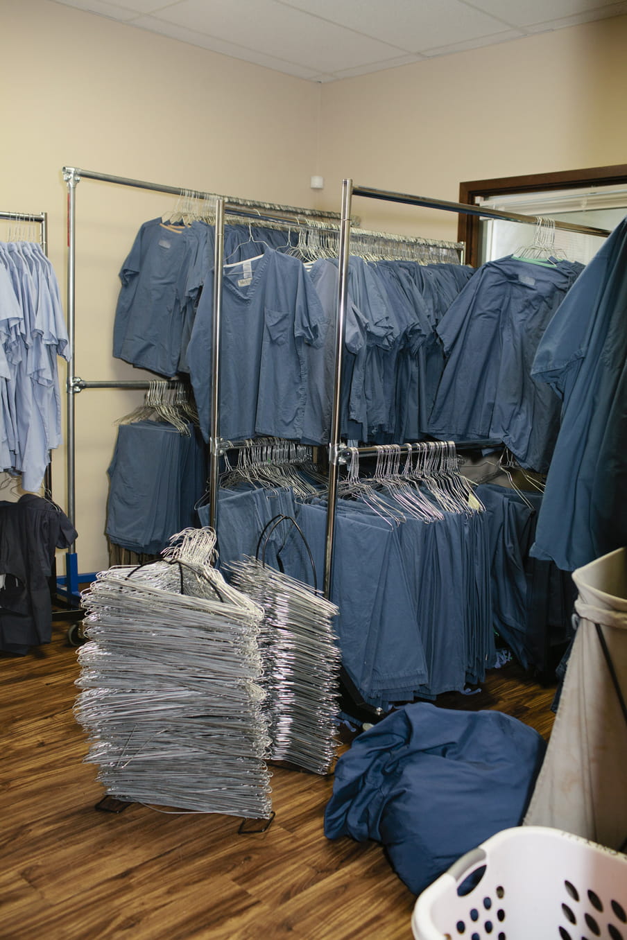 A photo taken in a room with four metal rails, filled with hanging blue smock uniforms worn by employees. At the front right, we see a white wash basket, a blue bag, and tall columns of silver metal hangers on the floor.