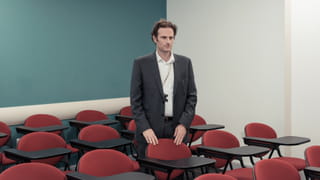 Man wearing a suit standing in a university classroom with his hands on one of the red chairs in it - the wall on the right is white, the one on the left is green.