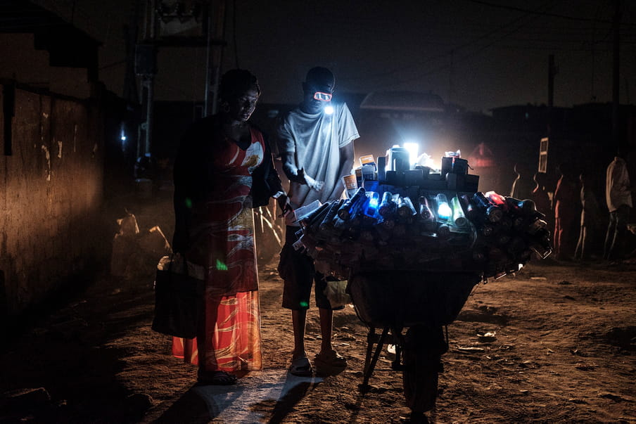 Photograph taken at night time in which a women with a long and light patterned orange dress stands next to a man wearing a white t-shirt and bermuda. The man has a lamp attached to the side of his head and he is showing her diverse flashlight options displayed on a structure on wheels. The ground is sandy. People are standing in line in the back. 