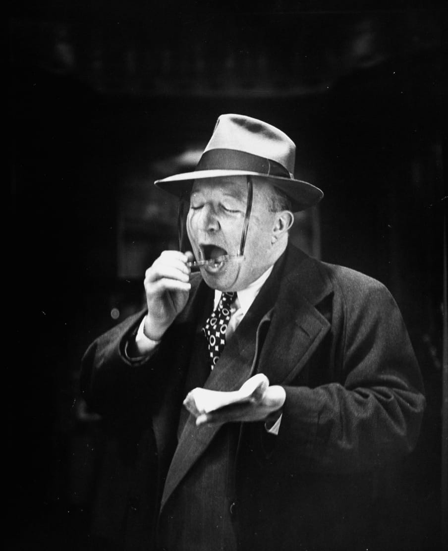 Old black and white photo of a man breathing on his glasses to clean them, in front of a mirror, shot from the back of the mirror