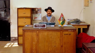 A Bolivian government worker with a black moustache sits behind a wooden desk. He's wearing a black hat and a light-blue shirt underneath a beige cardigan. A typewriter, a plate with his name and job function and a small Bolivian flag are placed in front of him. Some documents are lying on his desk. Behind him is a chest of lockable office drawers and another small table with a fax machine. A calendar is fastened to the wall. Two wooden chairs are visible on the side in front of the large desk.