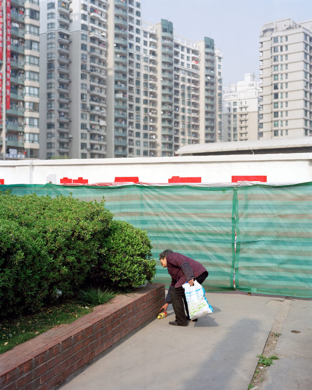 Photograph of a person picking up something that looks like a bottle from the road. In the background there are high rise buidlings