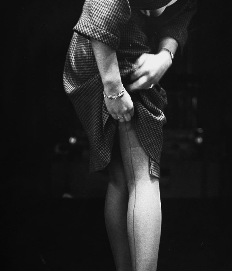 Old black and white photo of a woman checking her panty hoes in a mirror, shot from the back of the mirror