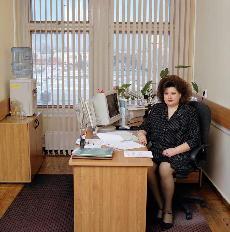 Russian secretary sits at a light-brown wooden desk. She has brown, shoulder-length hair in a perm. She wears a black skirt and a loose, black blouse with a fine, white pattern and black pumps. The desk is covered with some paperwork, a word processor, some stationery and plants. There’s a water dispenser and the office has brown-reddish carpet. The window’s white blinds are half-open and give way to a view of snow-covered landscape with some houses.