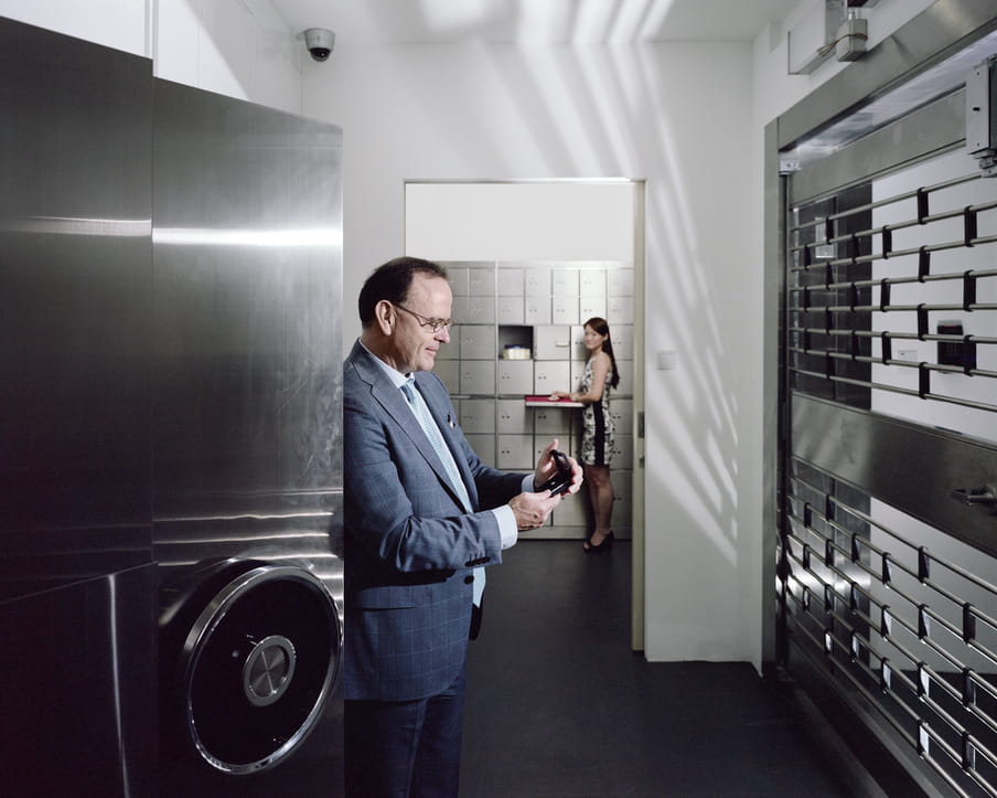 Man looking at a diamond while standing in front of a large safe. A woman standing in the back, looking at him.