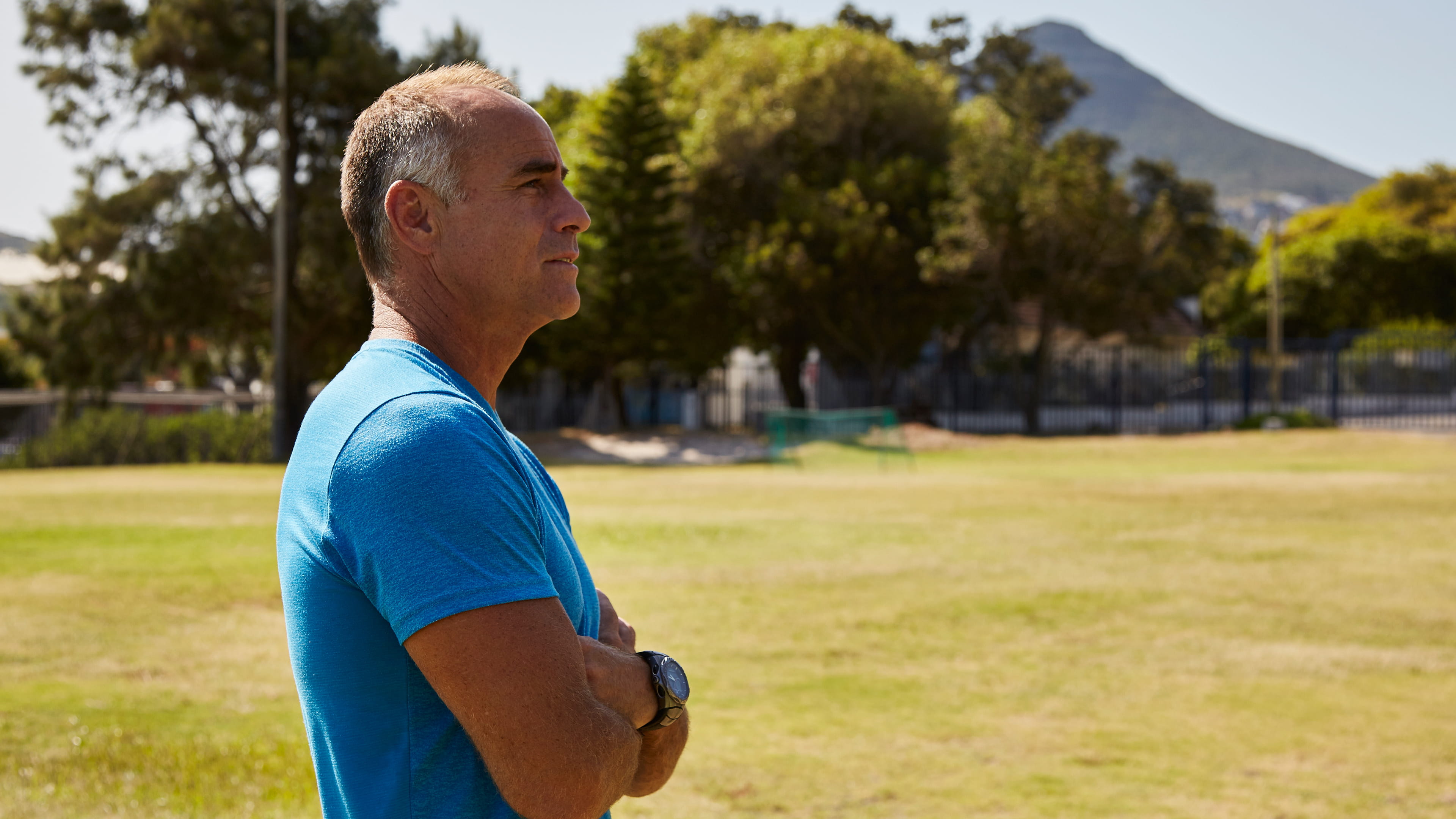 Photo of a man in a blue shirt in front of a green field, arms crossed and with trees and a mountain on the background