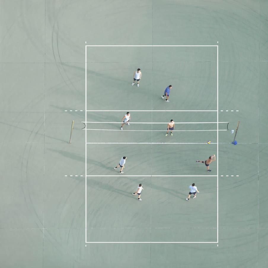 Photograph taken from above of men playing volleyball on a mint green court.