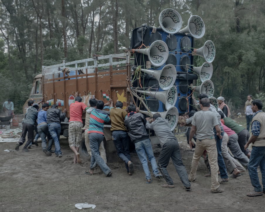 A group of men is pushing what seems to be a heavy truck filled with a elaborated sound system.