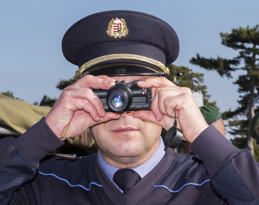 Picture showing a police man holding a camera-like surveillance device. The device is pointed towards the viewer as though he’s taking a picture. The man is wearing a police hat, a dark blue uniform jumper over a light blue shirt and a dark blue tie. The background includes a few military men, trees and blue sky.