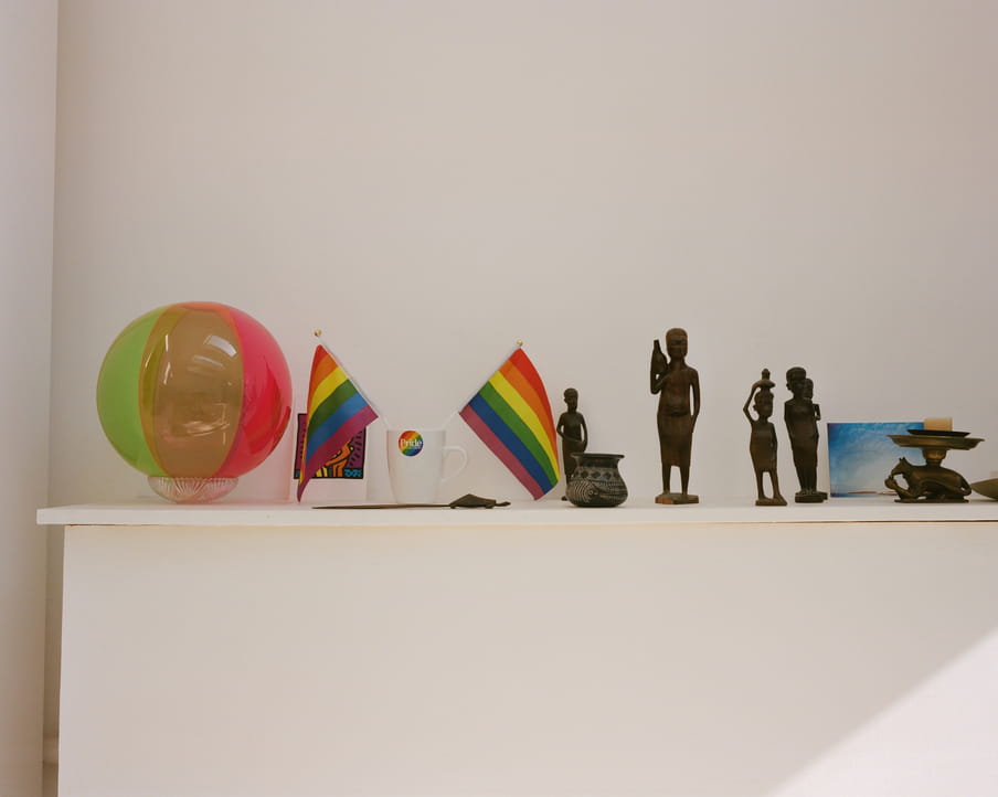 Picture of a white shelf against a white wall with various objects on it, left to right: inflatable small green and red beach ball; two rainbow flags sitting in a small white mug; five figurines of varying heights, looking like bronze or copper thin statues; and a blue postcard