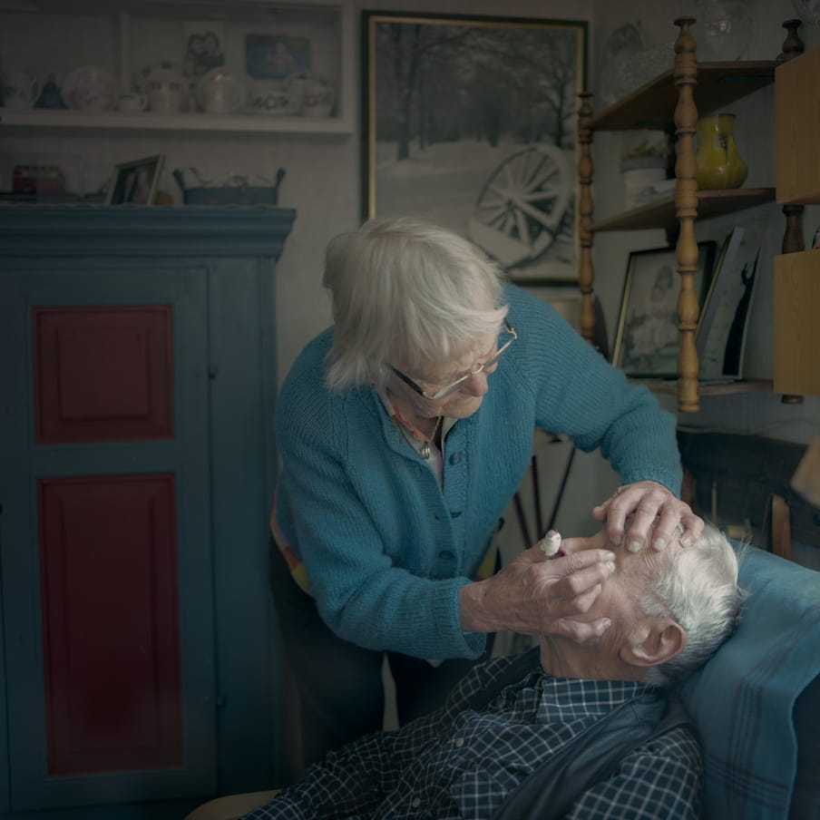 Photograph of an elderly couple. The man is sitting in a chair and the woman is bending over, appearing to get something out of the man his eye.