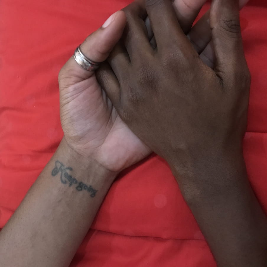 Two brown-skinned people hold hands in front of a red background. On one of their wrists there is a tattoo that reads ‘keep going’.