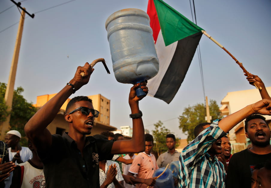 Photo of people on the street, front and centre showing a man hitting a large plastic bottle with a stick