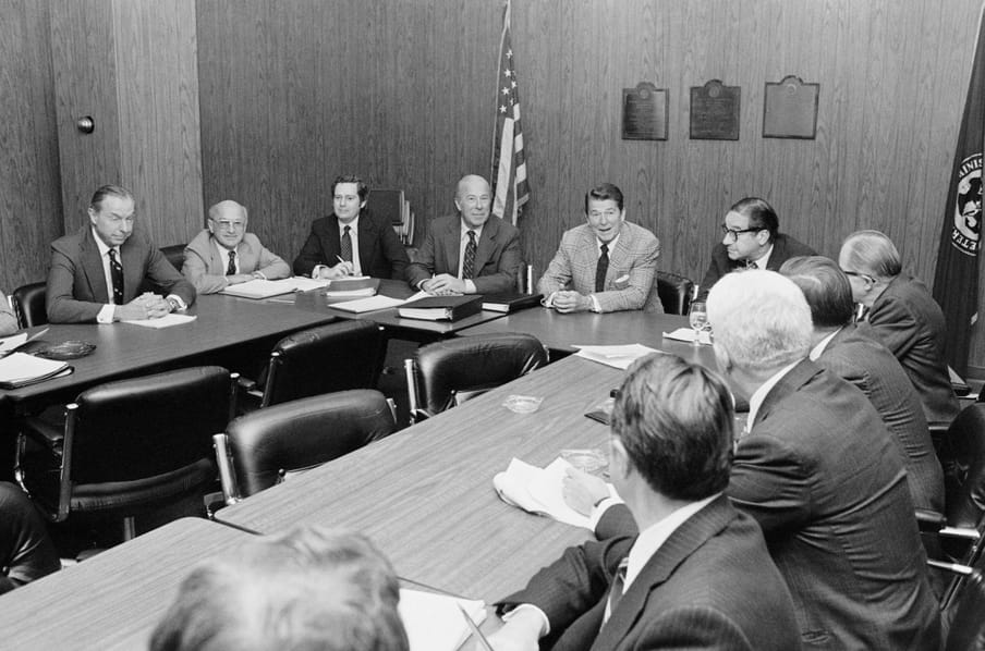 Table with 11 men in suits, american flag in the back