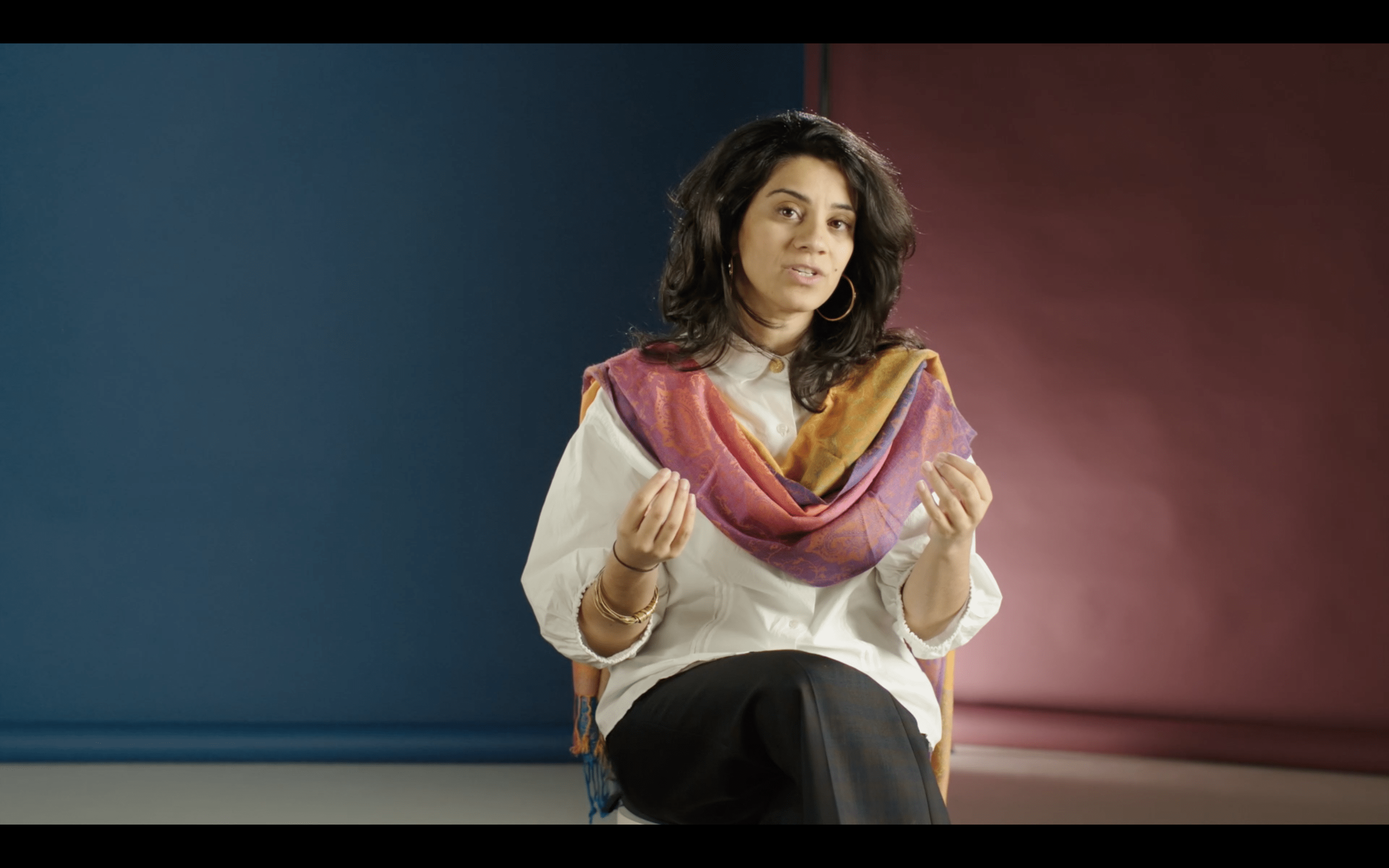 Screenshot of a moving image showing a woman talking towards the camera, a blue and pink background behind her. She’s wearing a colourful scarf and using her hands when she speaks