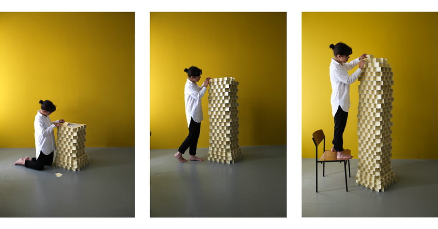 A series of three photographs showing a brown-haired women with glasses and a bun, in a large white shirt and black pants, barefoot, building a gigantic column with yellow playing cards. In the first image she is on her knees, she is standing on the second one and she is on a chair in the third one. The back wall is yellow and the floor if dark grey. 