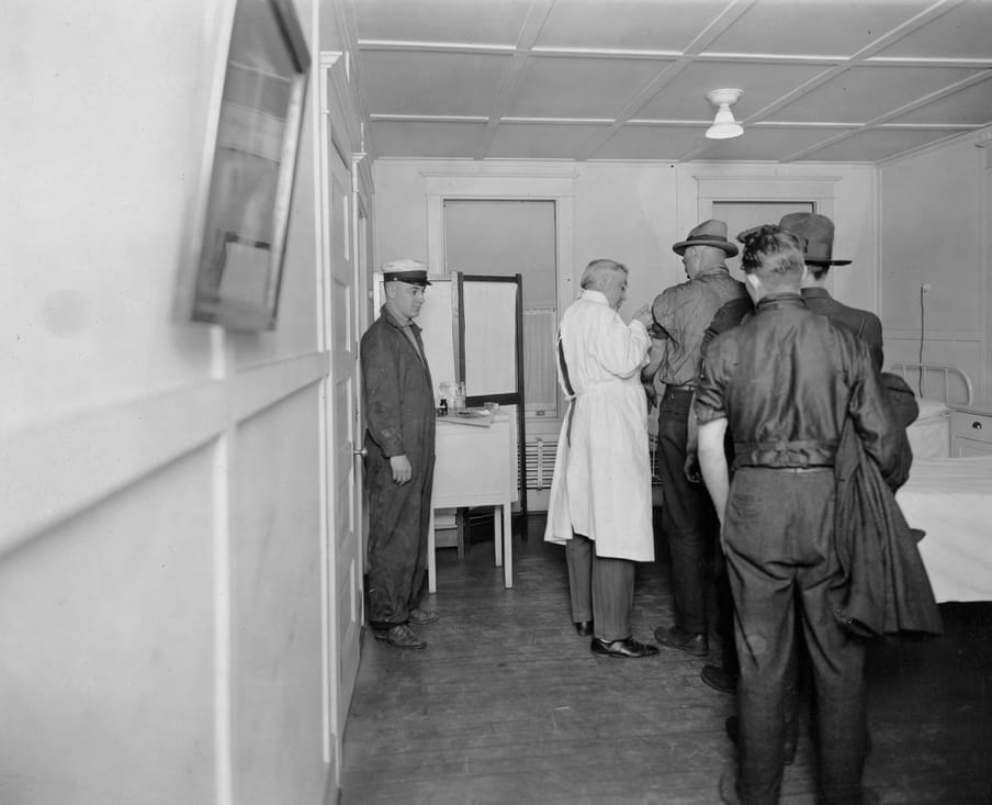 Black and white photograph of a group of men, in a line up, getting a shot in their left arm.