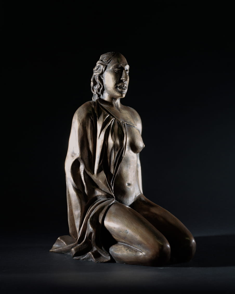 Photo of a bronze statue of a female figure sitting up straight on her knees, her mouth is open, showing teeth, she is wearing a cape - on a black background. Compared to the last image, you don’t see her giving birth in this one.