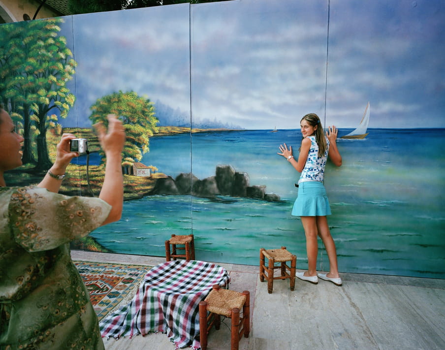 Just out of the frame, we see a man’s arm and side of his face as he takes a picture of a woman in a sleeveless blue and white top and turquoise summer mini skirt leaning with hands up against a triptych of paintings or a screen print of a calm sea, rocks and blue sky; she poses while looking backwards to the camera. In between the two people are stools and a low table with a tablecloth on it