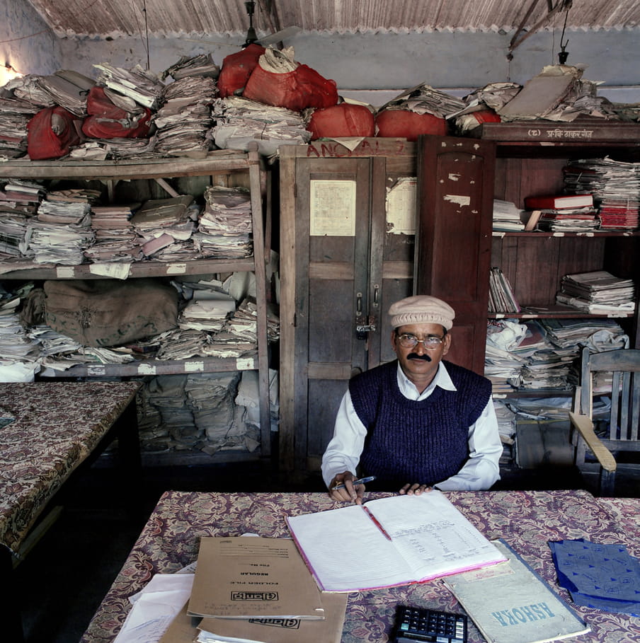 A male, Indian tax inspector sits at a desk covered with a floral print cover. A calculator and files with handwritten calculations are placed in front. He’s holding a pen and wears a white shirt, a beige cap and a dark-blue woollen vest. Behind him are shelves and cupboards bursting with files and sacks full of paperwork. The top of the cupboard is stacked with more paperwork, some of it in red sacks. They reach so high that they’re blocking the ventilator fan that’s fastened to the ceiling.