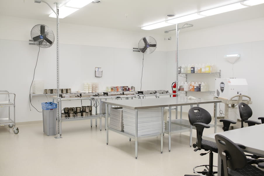 A large room with fluorescent lighting and silver metallic tables, with swivel chairs around a table in the foreground, small containers with different coloured liquids on shelving at the back, and two large fans hanging from brackets high on the walls.